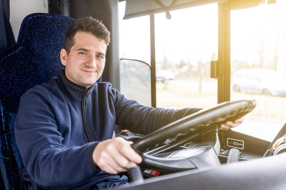 Ein Busfahrer in einer blauen Jacke sitzt lächelnd am Steuer, Sonnenlicht strömt herein, der Vereinsausflug kann beginnen